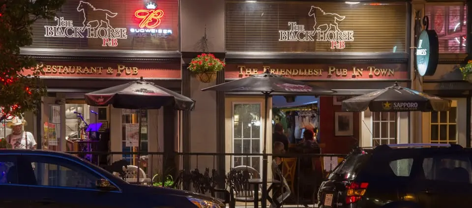 Night-time photo of the exterior of The Black Horse Pub in a downtown setting. The pub features dark signage with a horse graphic, a red Budweiser neon sign, and outdoor patio seating with black umbrellas. The windows are open, showing activity inside, and two cars are parked in front.