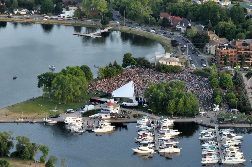 Aerial view of Del Crary Park hosting a large, crowded outdoor concert or event. The park is situated on a waterfront, featuring a stage with a distinctive white awning, and is surrounded by water docks full of parked boats. Residential areas, roads, and trees line the shore around the park.