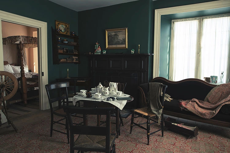Interior room of the Hutchison House Museum, likely a historical drawing-room or parlor. The walls are painted a deep teal or forest green. The room is furnished with a dark wooden table set for tea, a horsehair sofa, and features a dark wooden fireplace mantel. A doorway on the left opens to another room with a bed and a spinning wheel. Light streams in from a large window covered with sheer curtains on the right.