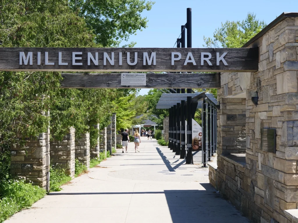 Query successful
The entrance to Millennium Park from King Street. A large, rustic wooden sign with raised white letters reading "MILLENNIUM PARK" spans the pathway. The entrance is framed by stone-clad columns and walls, with a sunny path leading into the park, where people can be seen walking.