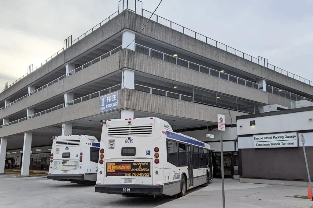 View of the Peterborough Bus Terminal (190 Simcoe St), showing the back of two Peterborough Transit buses parked in front of a multi-story, brutalist-style concrete parking garage. Signage indicates "Simcoe Street Parking Garage" and "Downtown Transit Terminal,"