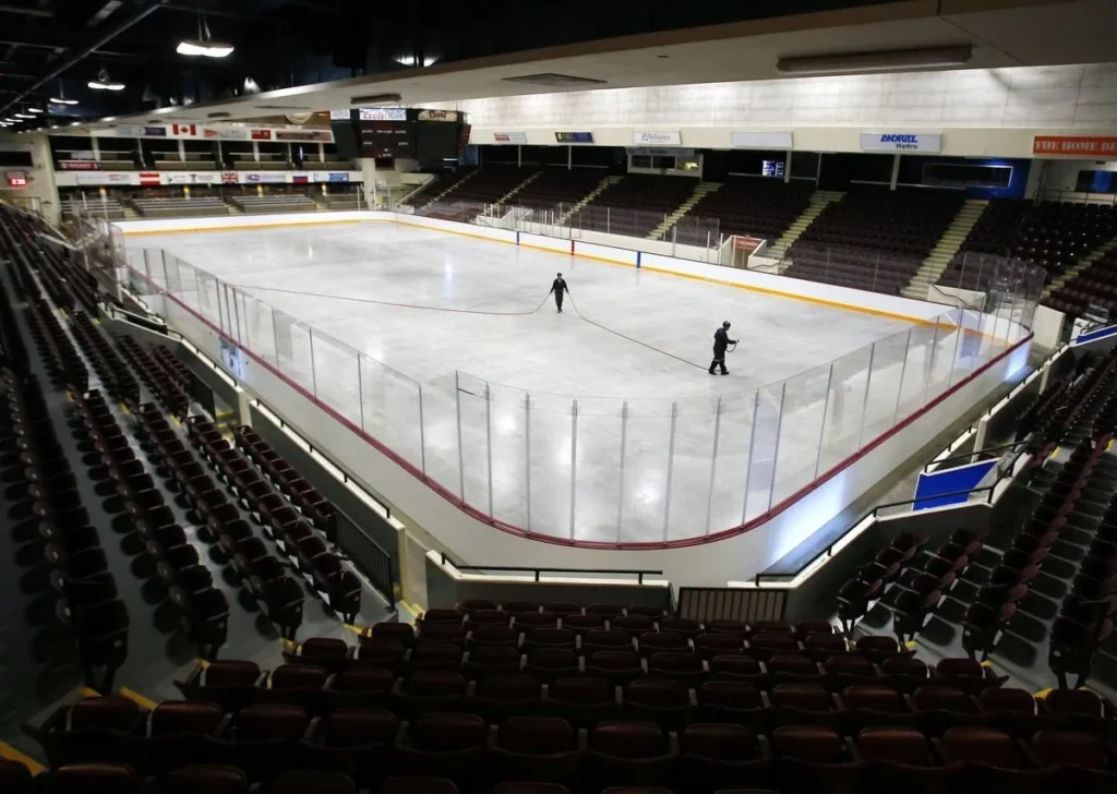 Query successful
Interior view of the Peterborough Memorial Centre (PMC) ice hockey arena. The ice surface is freshly maintained, surrounded by boards and glass. Two figures are visible on the ice, appearing to be workers. Dark seating fills the tiered stands surrounding the rink.
