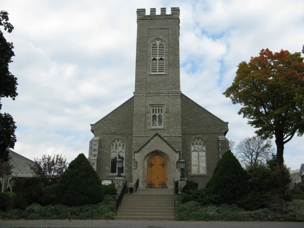 The front exterior of St. John the Evangelist Anglican Church in an autumn setting. The church is a stone structure featuring a prominent central square tower with crenellations, a pointed arch entrance with light wooden double doors, and Gothic-style windows. A set of stairs leads up to the entrance, flanked by manicured evergreen bushes.