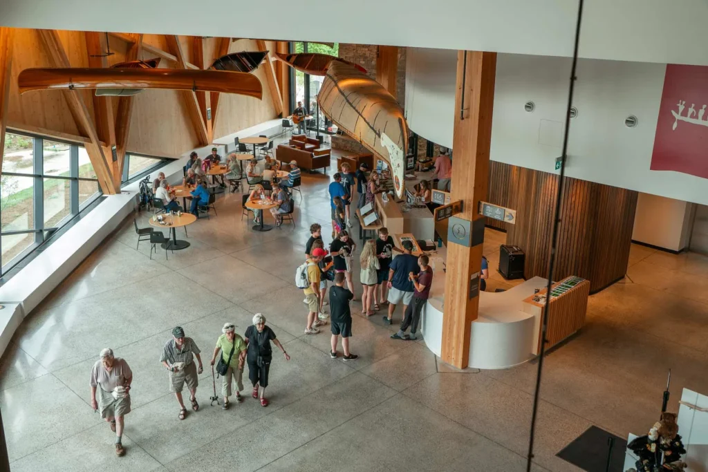 Overhead view of the bright, modern interior of The Canadian Canoe Museum. A large, historical canoe hangs dramatically from the high ceiling, suspended above a wooden reception desk and a crowd of visitors. Another canoe is mounted near large windows. People are seen exploring the space, lining up at a counter, and seated at small tables. The architecture features high ceilings, light-colored concrete floors, and exposed wooden structural elements.