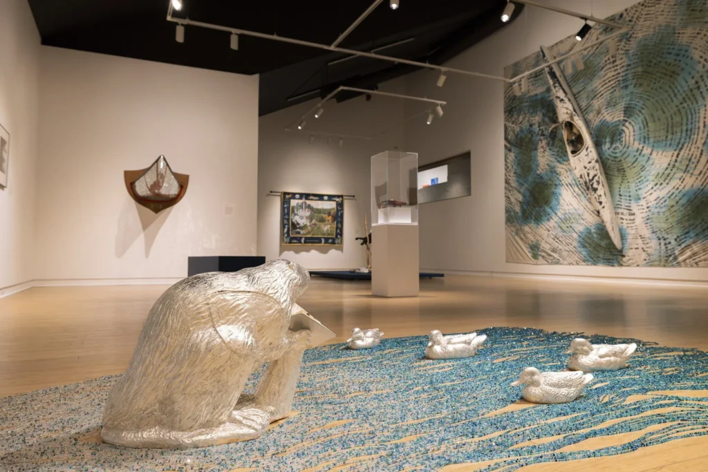 Query successful
Installation view of the exhibit "Watershed" at The Art Gallery of Peterborough. In the foreground, a large, crouching sculpture of an animal, seemingly covered in wrinkled silver foil, is on a floor installation of shimmering blue and tan materials, perhaps representing water and land. Several small, white duck sculptures are also on the 'water.' Various other artworks, including tapestries and a large canoe-themed piece, are visible on the white gallery walls in the background.