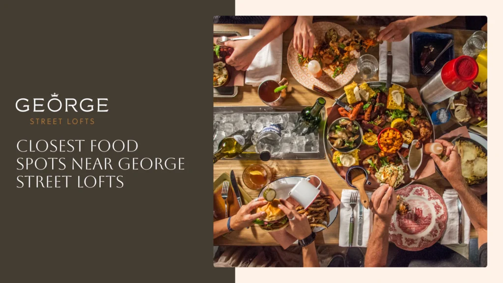 A top-down view shows a group of people sharing a large platter of southern-style food and drinks around a rustic wooden table. The image includes the George Street Lofts logo and the heading, "Closest Food Spots Near George Street Lofts."
