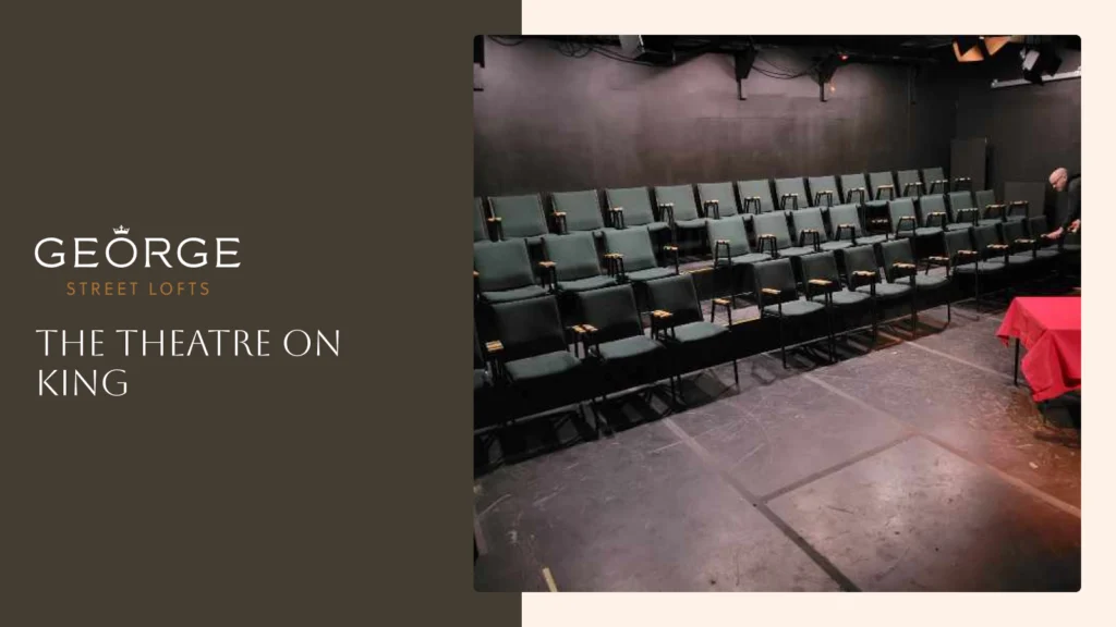 Interior view of The Theatre on King showing rows of dark green cushioned seats in a black-box theater setting, with the George Street Lofts logo on a side panel.